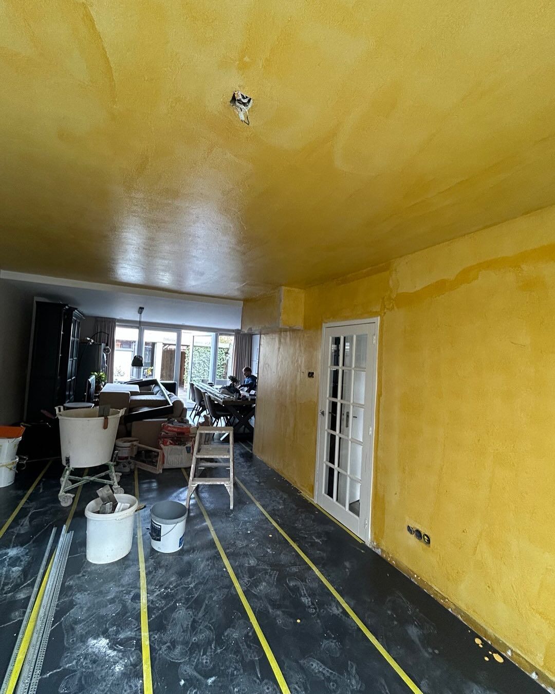 Lange woonkamer met gele voorstrijk op wanden en plafond, vloer afgeplakt
