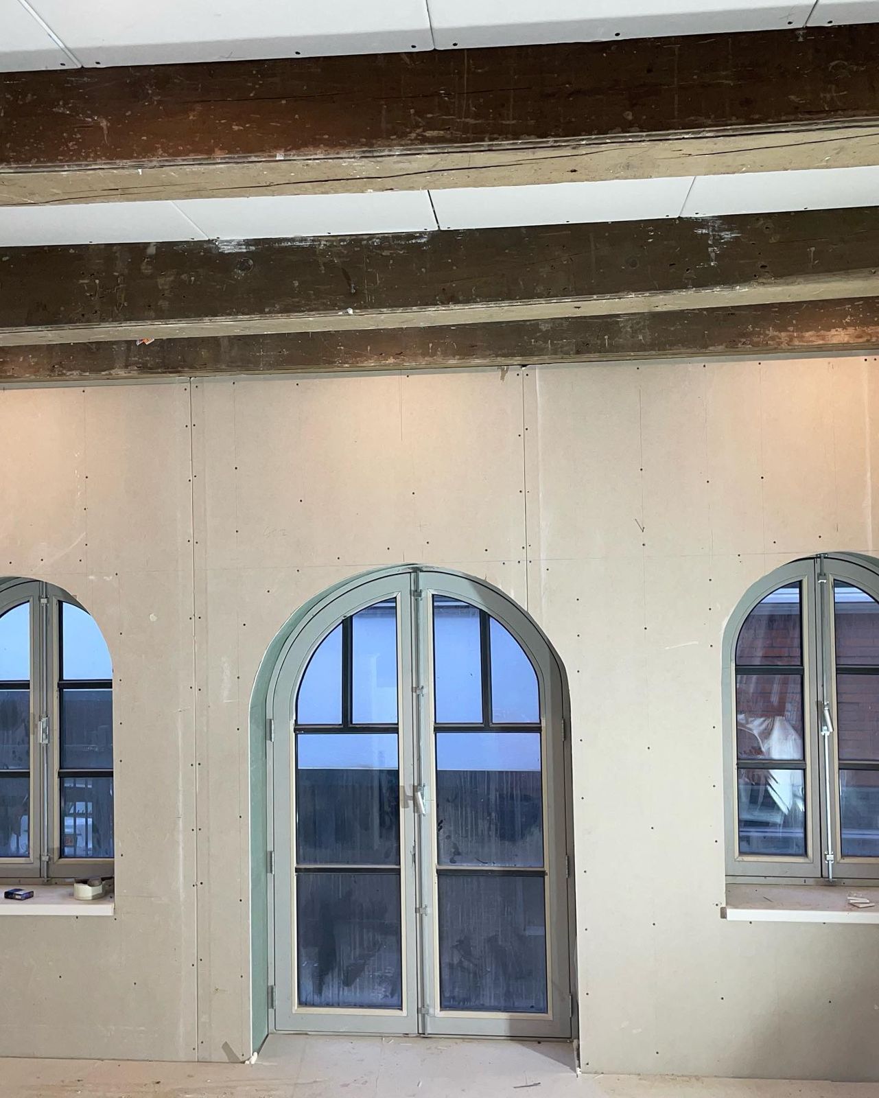 Wand van gipsplaten rond drie boogramen onder oude houten balken, klaar voor het stucwerk